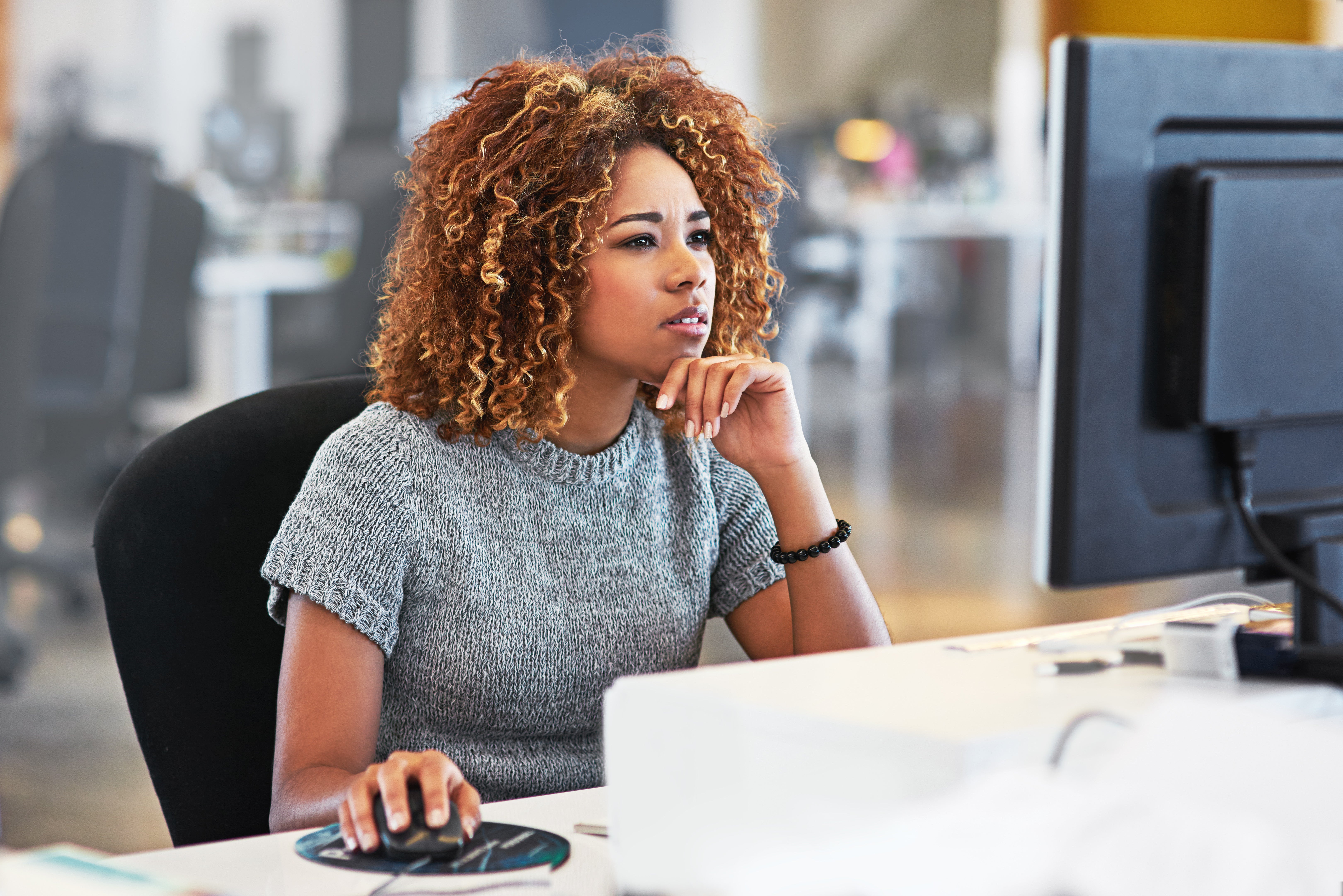 Woman looking at the screen of a desktop computer with her hand on a mouse and with a focused and pensive expression. 